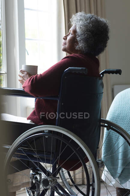 Mujer anciana afroamericana reflexiva sosteniendo la taza de café mirando por la ventana mientras está sentada en silla de ruedas en casa. permaneciendo en casa en aislamiento en cuarentena - foto de stock