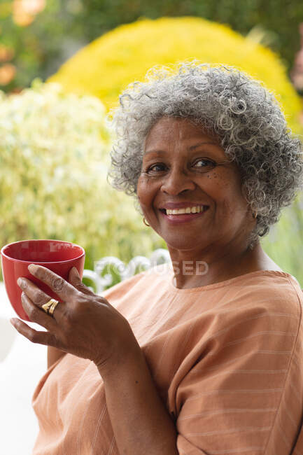 Портрет африканской пожилой женщины с кофейной чашкой, улыбающейся сидя на крыльце. оставаться в изоляции в карантинной изоляции — стоковое фото