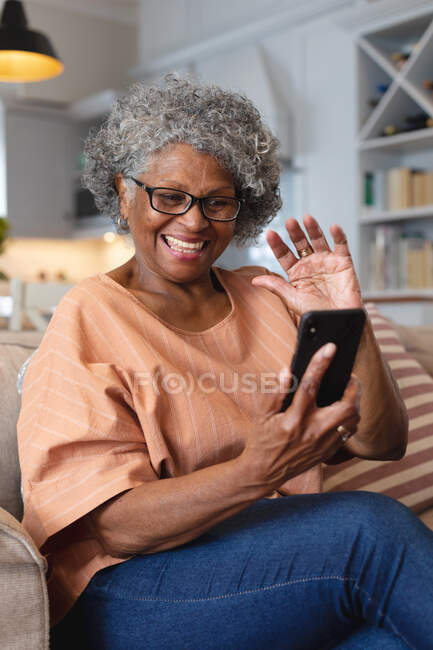Donna anziana afroamericana che sorride e saluta mentre riceve una videochiamata sullo smartphone a casa. stare a casa in isolamento personale in quarantena — Foto stock