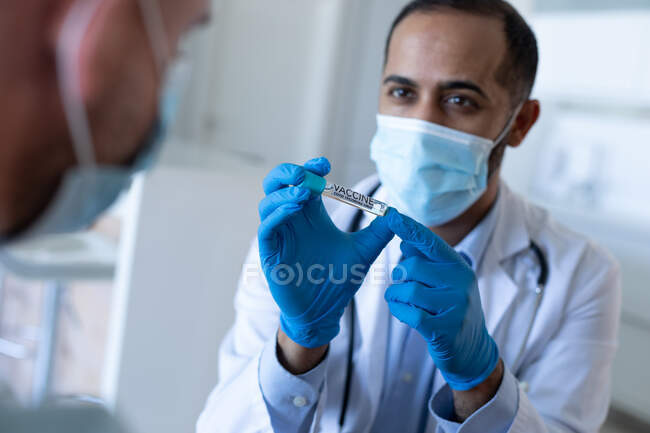 Médico masculino de raza mixta que usa mascarilla facial preparando la vacuna para el paciente masculino. higiene protección sanitaria durante la pandemia del coronavirus covid 19. - foto de stock
