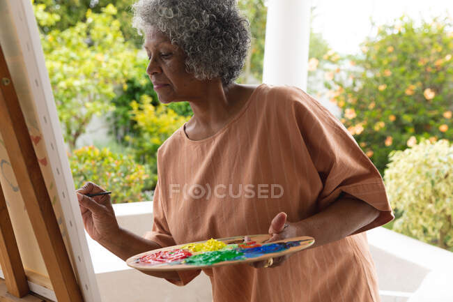 Задумчивая африканская старшая женщина рисует на холсте, стоя на крыльце дома. оставаться в изоляции в карантинной изоляции — стоковое фото
