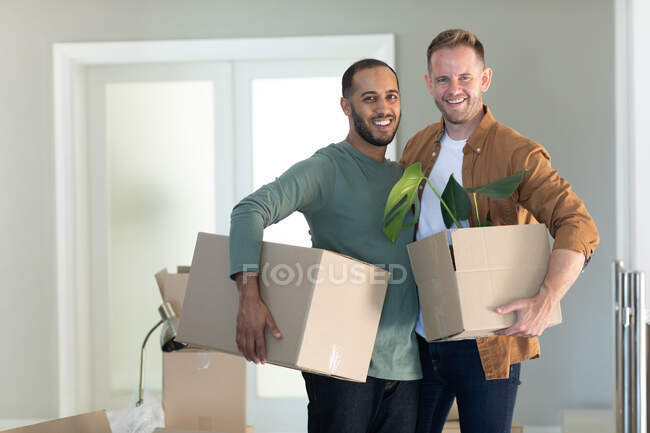 Multi étnico gay masculino casal segurando caixas olhando para câmera e sorrindo em casa. aproveitando o tempo em casa em auto-isolamento durante o bloqueio de quarentena. — Fotografia de Stock