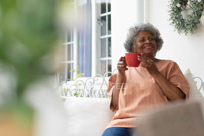 Mujer mayor afroamericana sosteniendo taza de café sonriendo mientras está sentada en el porche de la casa. permaneciendo en aislamiento en cuarentena - foto de stock