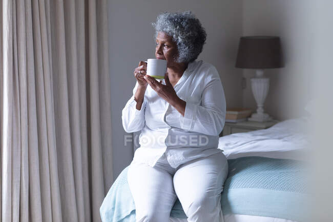 Thoughtful african american senior woman drinking coffee while sitting on bed at home. staying at home in self isolation in quarantine lockdown — Stock Photo