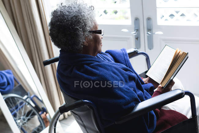 Mujer mayor afroamericana leyendo un libro mientras está sentada en silla de ruedas en casa. permaneciendo en casa en aislamiento en cuarentena - foto de stock