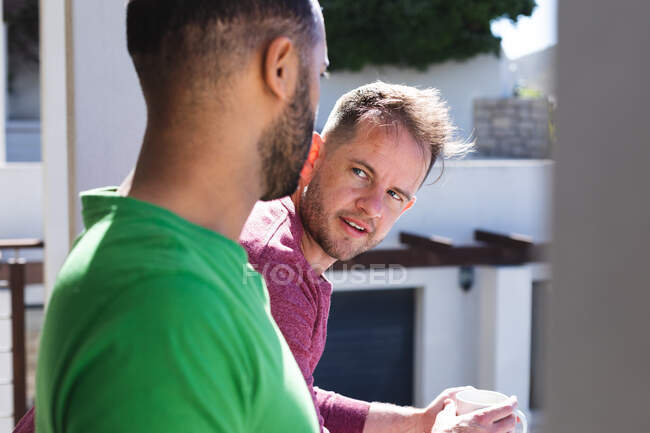 Casal masculino gay multi étnico falando na varanda ao sol. ficar em casa em auto-isolamento durante o confinamento de quarentena. — Fotografia de Stock