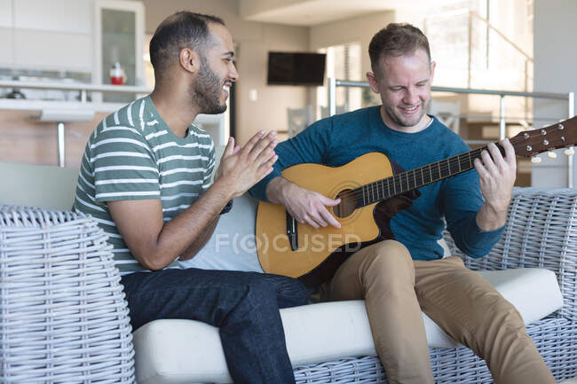 Couple masculin gay multi ethnique assis sur le canapé un jouer de la guitare. rester à la maison en isolement personnel pendant le confinement en quarantaine. — Photo de stock
