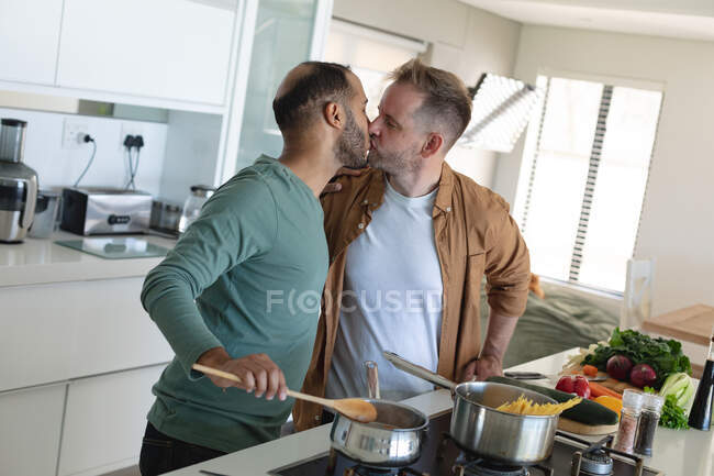 Multi ethnic gay male couple preparing food together and kissing at home. staying at home in self isolation during quarantine lockdown. — Stock Photo