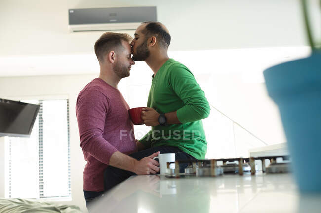 Multi ethnic gay male couple sitting in kitchen drinking coffee and kissing at home. Staying at home in self isolation during quarantine lockdown. — Stock Photo