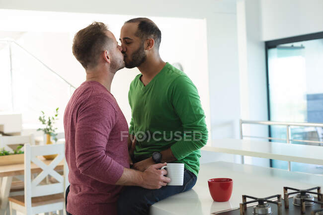 Couple masculin gay multi ethnique assis dans la cuisine buvant du café et s'embrassant à la maison. Rester à la maison en isolement personnel pendant le confinement en quarantaine. — Photo de stock