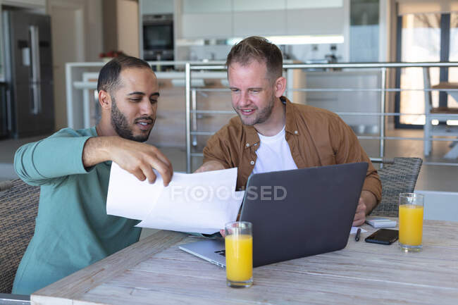 Multi ethnic gay male couple going through bills and using laptop at home. staying at home in self isolation during quarantine lockdown. — Stock Photo