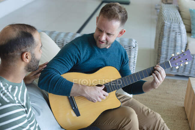 Multi casal masculino gay étnico sentado no sofá um tocando guitarra. ficar em casa em auto-isolamento durante o confinamento de quarentena. — Fotografia de Stock