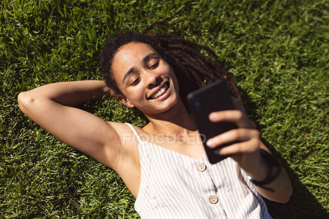 Lächelnde Afroamerikanerin im Gras liegend und mit Smartphone im Park. Digitaler Nomade unterwegs. — Stockfoto