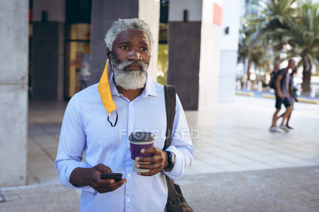 African american senior man wearing hanging face mask walking in street holding coffee and smartphone. digital nomad out and about in the city during coronavirus covid 19 pandemic. — Stock Photo