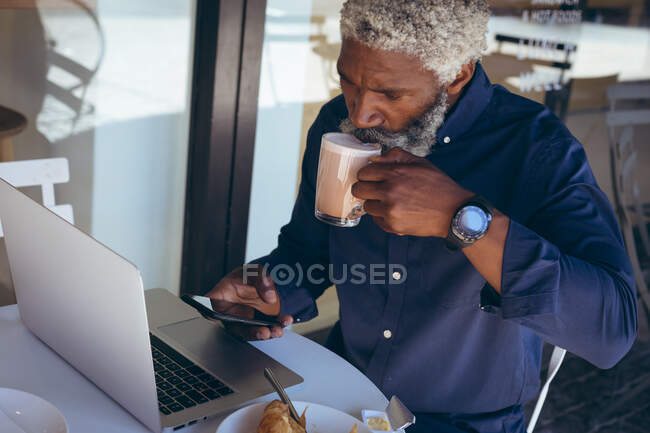 Ein älterer afroamerikanischer Mann sitzt am Tisch vor einem Café und trinkt Kaffee mit Laptop und Smartphone. digitaler Nomade in der Stadt unterwegs. — Stockfoto