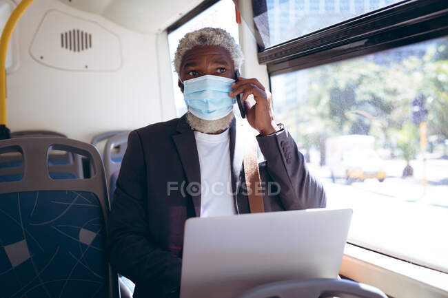 Afro-americano sênior vestindo máscara facial sentado no ônibus usando laptop falando no smartphone. nômade digital para fora e sobre na cidade durante coronavírus covid 19 pandemia. — Fotografia de Stock