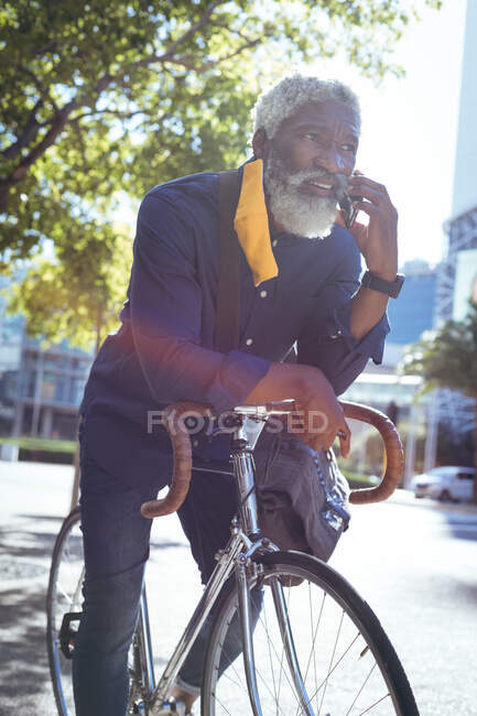 Hombre mayor afroamericano con máscara facial apoyado en la bicicleta en la calle hablando en el teléfono inteligente. nómada digital en la ciudad durante la pandemia de coronavirus covid 19. - foto de stock