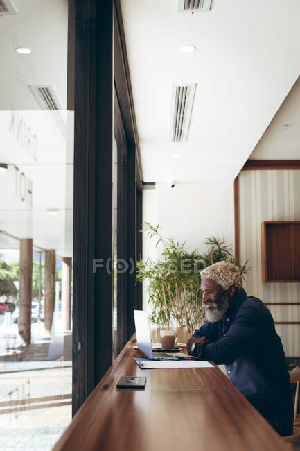 Uomo anziano afroamericano seduto a tavola in un bar a lavorare con il computer portatile e sorridente. nomade digitale in giro per la città. — Foto stock