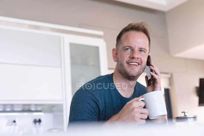 Белый мужчина улыбается, пьет кофе и разговаривает дома со смартфоном. Оставаться дома в изоляции во время карантинной изоляции. — стоковое фото
