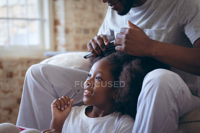 African american man sitting on bed doing his daughter hair. staying at home in self isolation during quarantine lockdown. — Stock Photo