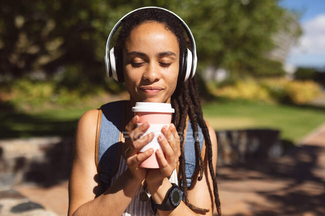 Mujer afroamericana sonriente con auriculares sentados bebiendo café en el parque. Nómada digital sobre la marcha estilo de vida. - foto de stock