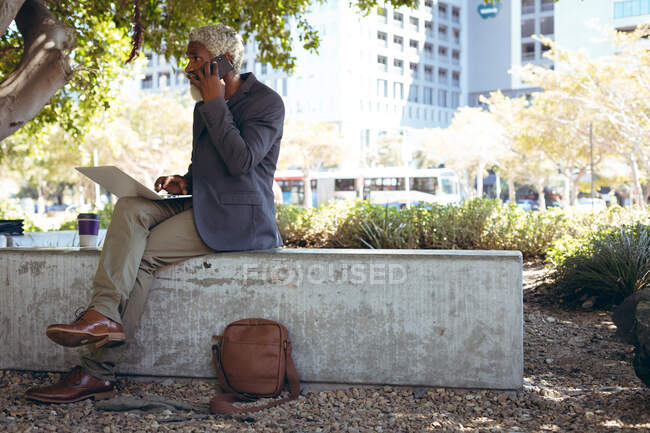 Hombre mayor afroamericano sentado en la pared en la calle usando un ordenador portátil. nómada digital en la ciudad. - foto de stock