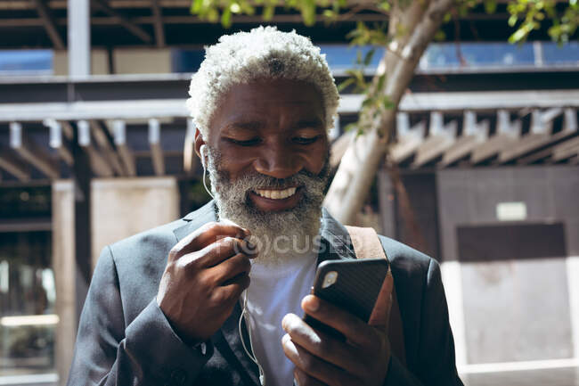Африканский пожилой человек в наушниках стоит на улице, используя смартфон и улыбаясь. цифровая реклама в городе. — стоковое фото