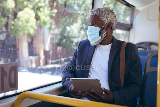 Uomo anziano afroamericano con maschera facciale seduto sull'autobus con tablet digitale che guarda fuori dalla finestra. nomade digitale in giro per la città durante coronavirus covid 19 pandemia. — Foto stock