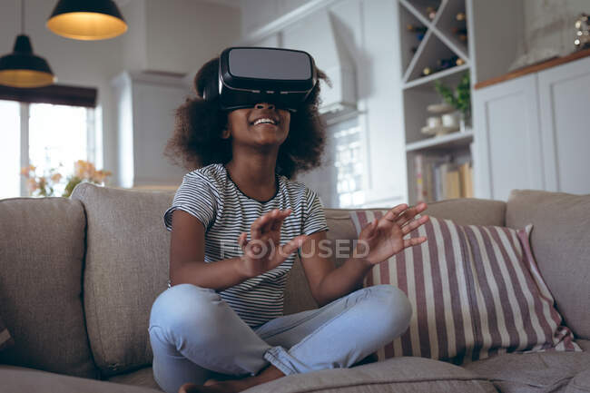 Chica afroamericana sentada en la sala de estar usando auriculares vr. permanecer en casa en aislamiento durante el bloqueo de cuarentena. - foto de stock