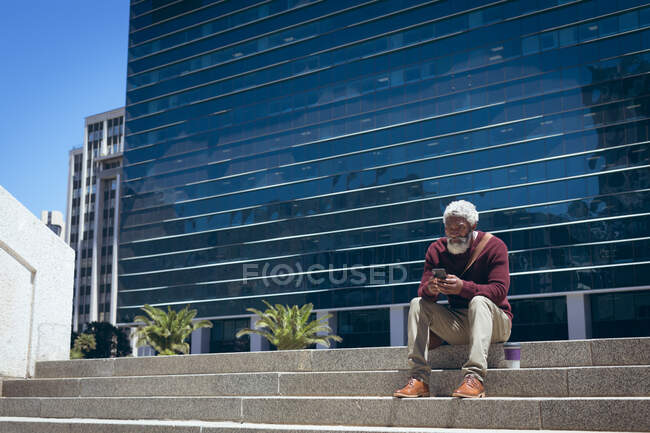 Hombre mayor afroamericano sentado en escalones fuera del edificio con café para llevar usando un teléfono inteligente. nómada digital en la ciudad. - foto de stock