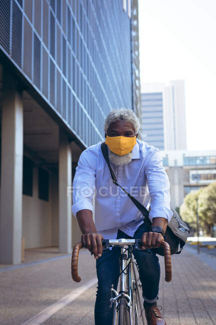 Старший африканский американец в маске для лица катается на велосипеде по улице. Цифровой кочевник в городе во время пандемии коронавируса. — стоковое фото