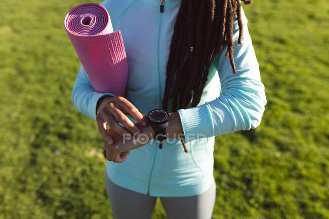 В середині афроамериканського парку жінка, яка займається фізичними вправами, користується смартвичем і несе мату йоги. Здоровий зовнішній спосіб життя. — стокове фото