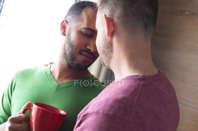 Couple masculin gay multi ethnique prenant un café et se câlinant à la maison. Rester à la maison en isolement personnel pendant le confinement en quarantaine. — Photo de stock