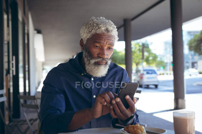 Afrikanisch-amerikanischer Senior sitzt mit Smartphone am Tisch vor einem Café und trinkt Kaffee. digitaler Nomade in der Stadt unterwegs. — Stockfoto