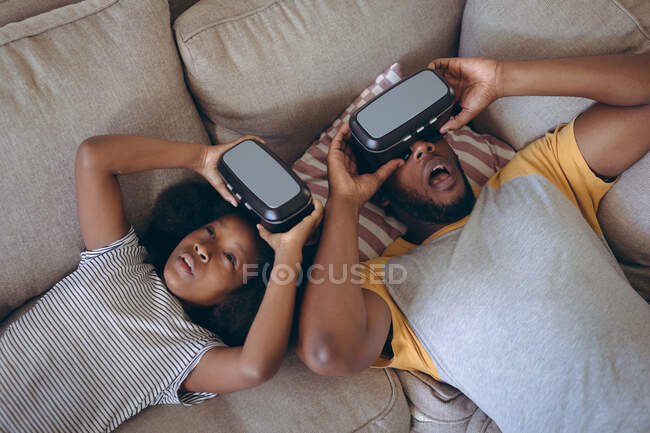 Homme afro-américain et sa fille couchés dans le salon en utilisant un casque vr. rester à la maison en isolement personnel pendant le confinement en quarantaine. — Photo de stock