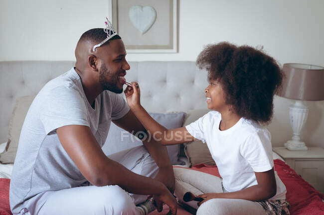 African american man wearing tiara having makeup put on by his daughter. staying at home in self isolation during quarantine lockdown. — Stock Photo