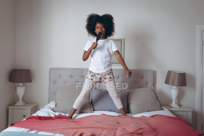 Chica afroamericana de pie en la cama sosteniendo cepillo de pelo fingiendo cantar. permanecer en casa en aislamiento durante el bloqueo de cuarentena. - foto de stock