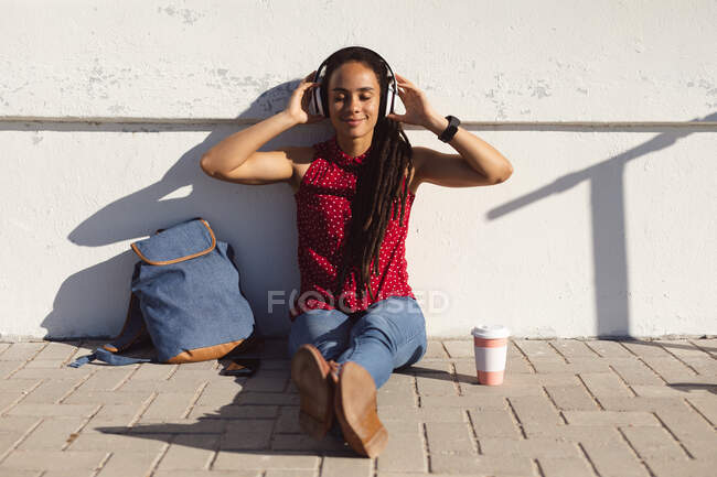 Feliz mulher afro-americana sorrindo com os olhos fechados, sentada no pavimento e ouvindo música. Nômade digital em movimento estilo de vida. — Fotografia de Stock