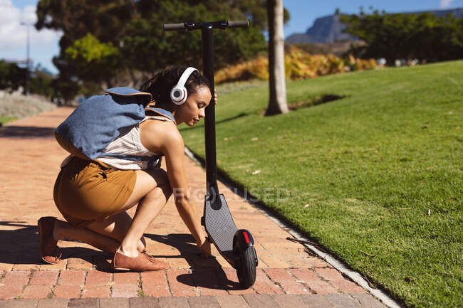 Donna afroamericana con le cuffie che parcheggia uno scooter in strada. Nomade digitale in movimento lifestyle. — Foto stock