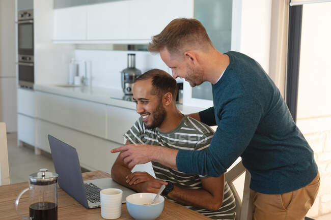 Casal masculino gay multi étnico sorrindo e comendo café da manhã e usando laptop em casa. Ficar em casa em auto-isolamento durante o bloqueio de quarentena. — Fotografia de Stock