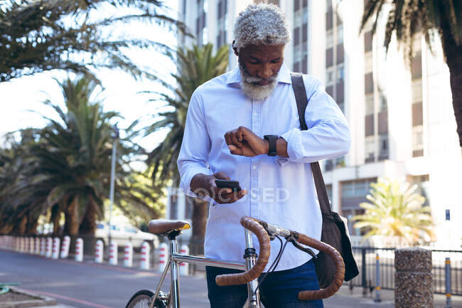 Homem sênior afro-americano de pé na rua com bicicleta segurando smartphone e verificando relógio. nômade digital para fora e sobre na cidade. — Fotografia de Stock