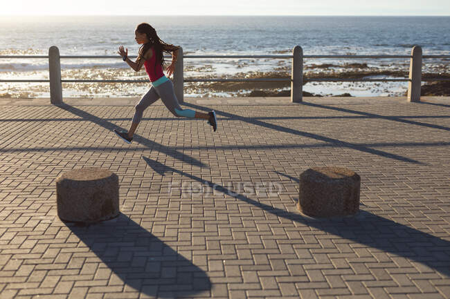 African american woman concentrating, exercising on a promenade by the sea running. Fitness healthy outdoor lifestyle. — Stock Photo