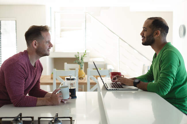 Multi ethnic gay male couple smiling and sitting in kitchen drinking coffee and using laptop at home. Staying at home in self isolation during quarantine lockdown. — Stock Photo