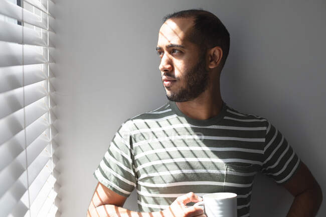 Homme de race mixte debout près de la fenêtre pensant, prenant un café à la maison. Rester à la maison en isolement personnel pendant le confinement en quarantaine. — Photo de stock