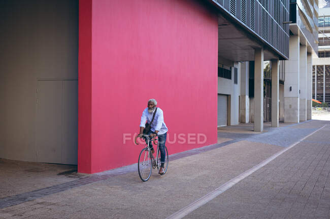 Uomo anziano afroamericano in bicicletta per strada oltre il muro rosso. nomade digitale in giro per la città. — Foto stock