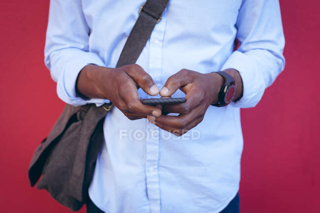 Sección media del hombre de pie contra la pared roja en la calle con el teléfono inteligente. nómada digital en la ciudad. - foto de stock