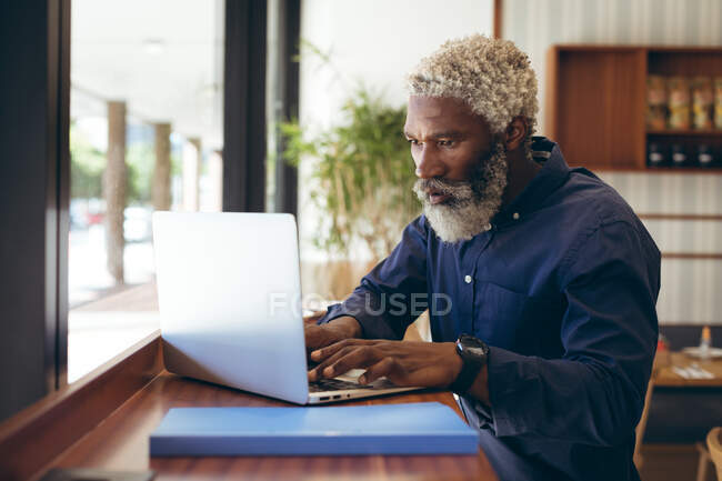 Hombre mayor afroamericano sentado en la mesa en la cafetería trabajando con un portátil. nómada digital en la ciudad. - foto de stock