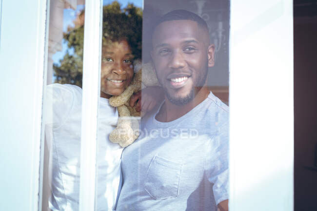 Afroamerikanerin und ihr Vater halten einen Teddybär in der Hand, der durch das Fenster schaut. Während der Quarantäne zu Hause bleiben und sich selbst isolieren. — Stockfoto