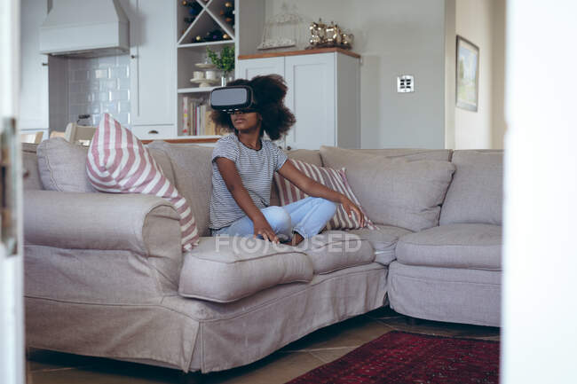 Afroamerikanerin sitzt im Wohnzimmer mit vr headset. Während der Quarantäne zu Hause bleiben und sich selbst isolieren. — Stockfoto