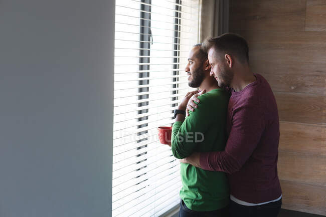Multi étnico gay casal masculino beber café, sorrindo e abraçando em casa. Ficar em casa em auto-isolamento durante o bloqueio de quarentena. — Fotografia de Stock